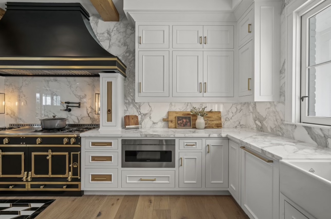 Elegant kitchen design with white cabinetry, brass hardware, and Calacatta marble countertops and backsplash, paired with a black-and-gold range hood.