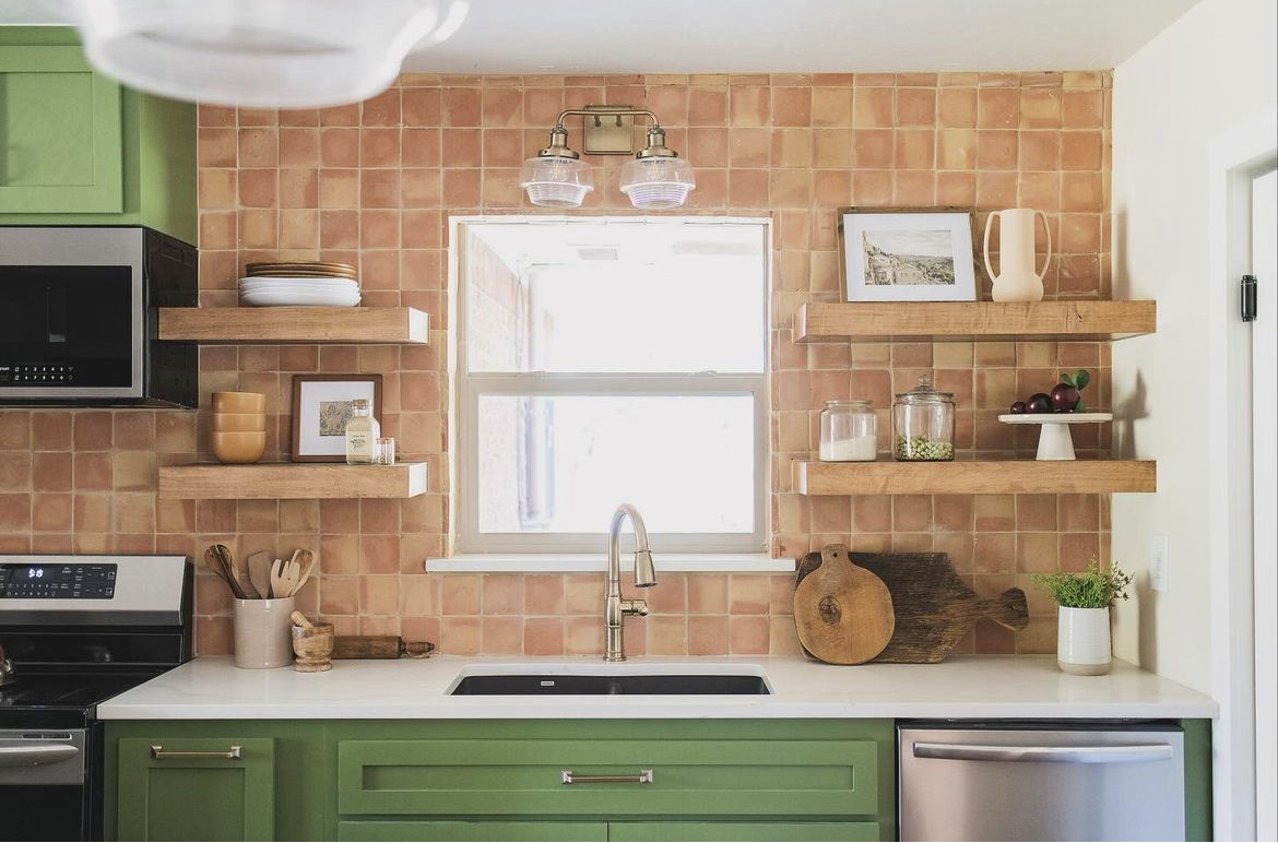 Modern kitchen with green cabinets, white countertops, terra cotta tile backsplash, and floating wooden shelves.